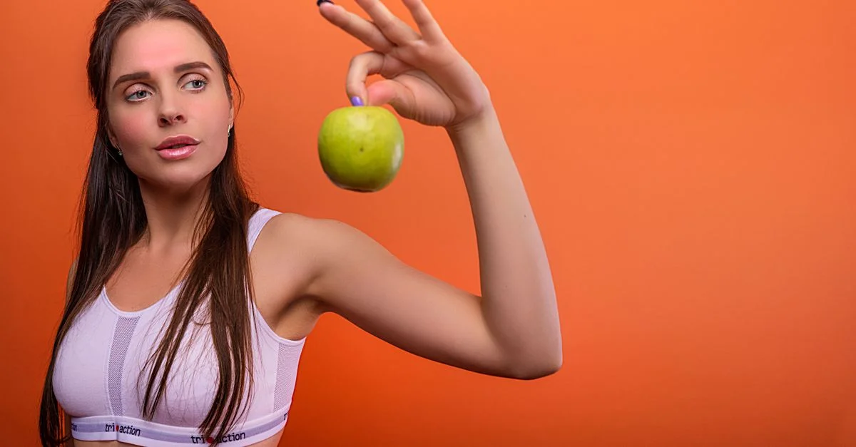 Joven mujer en ropa deportiva sosteniendo una manzana verde frente a un fondo naranja.
