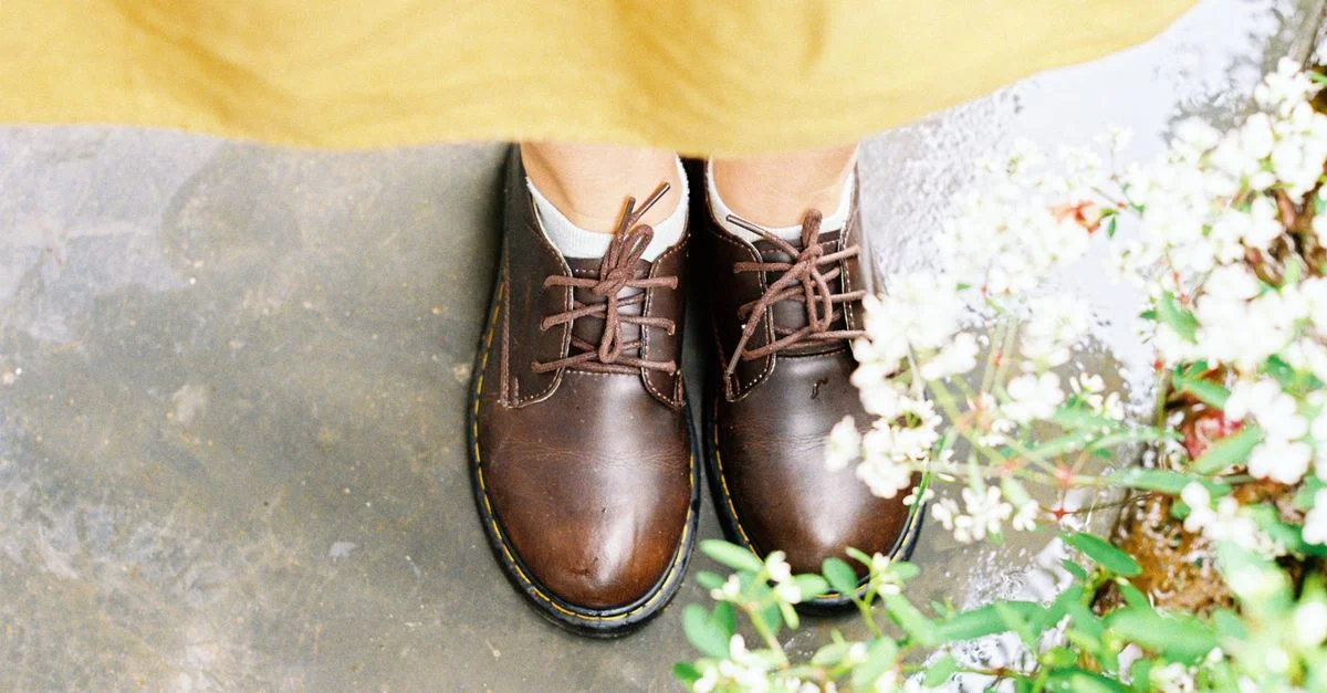 Acercamiento de zapatos de cuero marrón en suelo mojado, rodeados de pequeñas plantas.