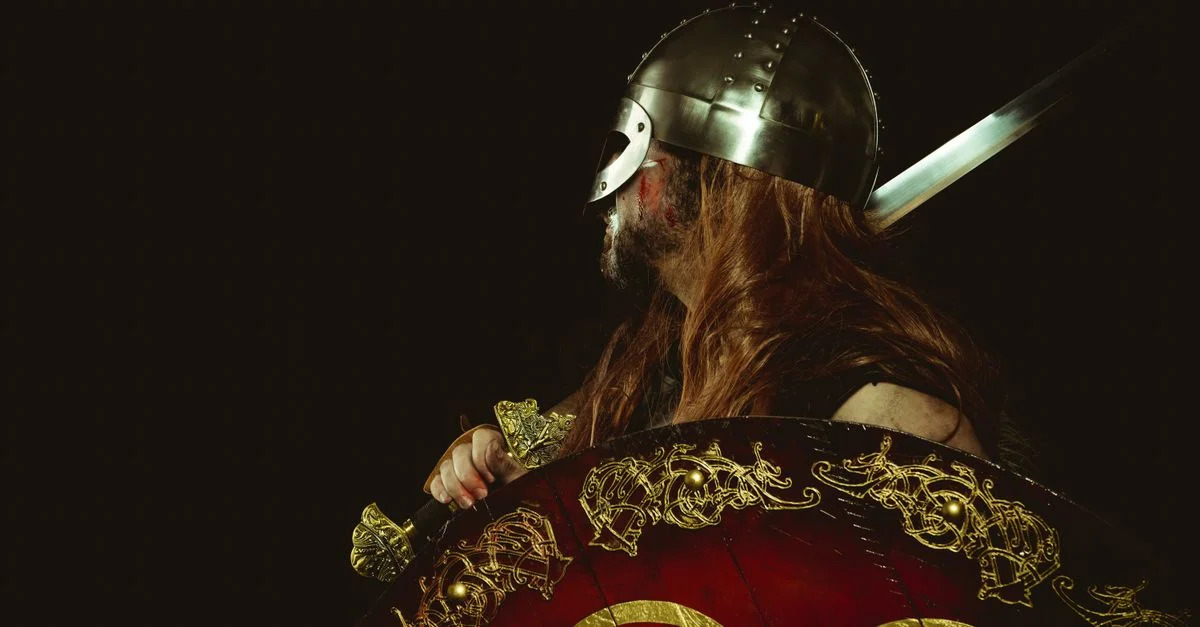 Perfil lateral de un guerrero vikingo con armadura tradicional, sosteniendo un escudo decorado y una espada.