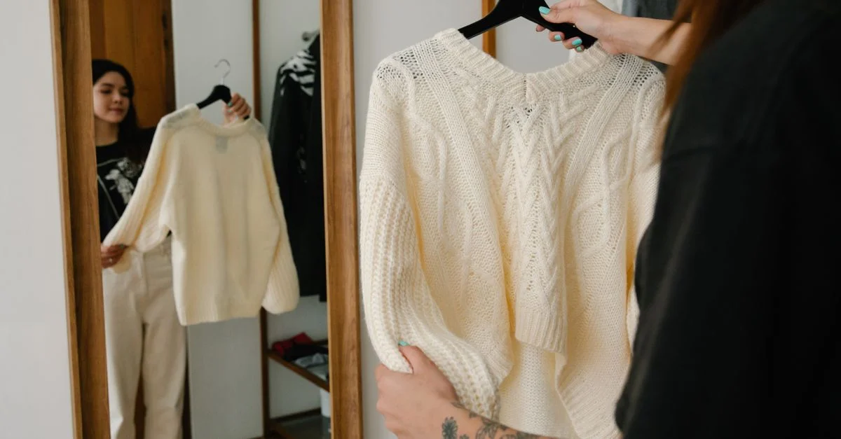 Mujer sosteniendo y probándose un suéter de punto mientras observa su reflejo en un espejo, mostrando estilo de moda.